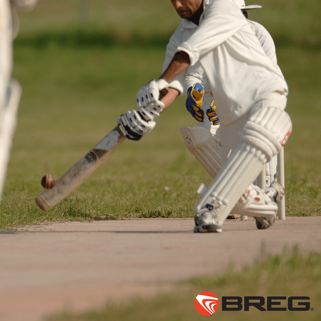 Cricket batter in white gear hitting ball on grassy pitch with Breg logo for cold therapy devices