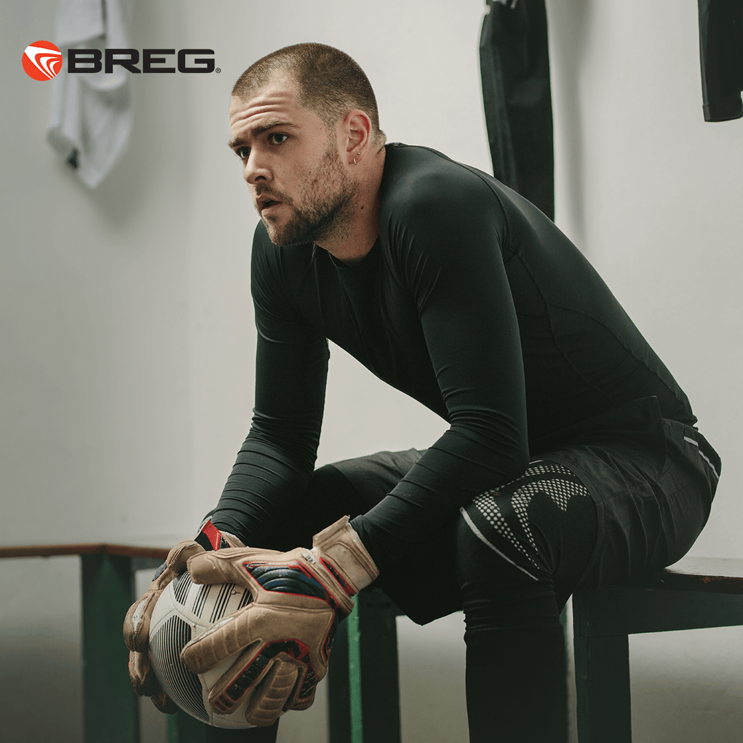 Focused male soccer goalkeeper in black athletic wear holding a ball, sitting on locker room bench