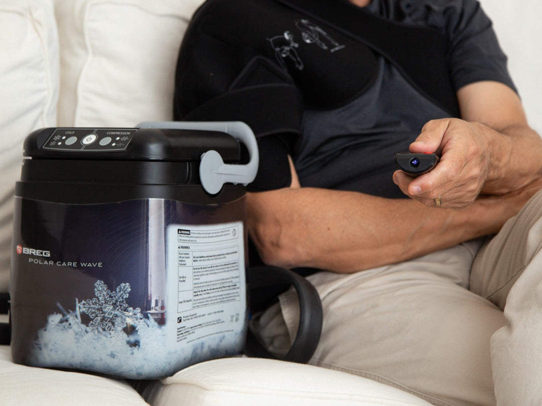 Person using Breg Polar Care Wave cold therapy device with remote control for arm injury recovery on white couch